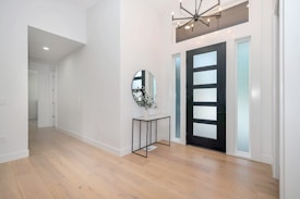 A modern hallway with light wood flooring and white walls. On the right, there is a black front door with frosted glass panels, flanked by two tall frosted windows. Above the door is a horizontal window. A contemporary black chandelier hangs from the ceiling. On the left side, a small black metal console table holds a round mirror and a vase with green foliage.