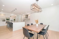 A modern kitchen and dining area featuring a spacious island with high black chairs, pendant lighting, and sleek white cabinetry. The dining table is wooden with black chairs and a vase with dried flowers. Neutral tones create a minimalist and clean aesthetic.