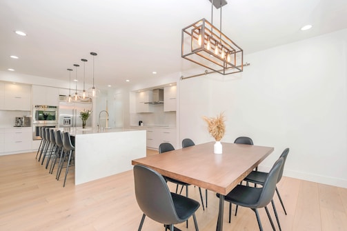 A modern kitchen and dining area featuring a spacious island with high black chairs, pendant lighting, and sleek white cabinetry. The dining table is wooden with black chairs and a vase with dried flowers. Neutral tones create a minimalist and clean aesthetic.
