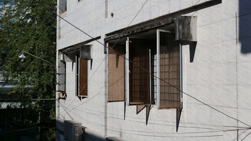 A building facade with three open windows featuring metal grills. The walls are tiled with flat, white panels. A part of a tree with green foliage is visible on the left side. Numerous black wires crisscross in front of the building, and an air conditioning unit is mounted beneath one of the windows.