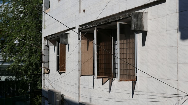 A building facade with three open windows featuring metal grills. The walls are tiled with flat, white panels. A part of a tree with green foliage is visible on the left side. Numerous black wires crisscross in front of the building, and an air conditioning unit is mounted beneath one of the windows.