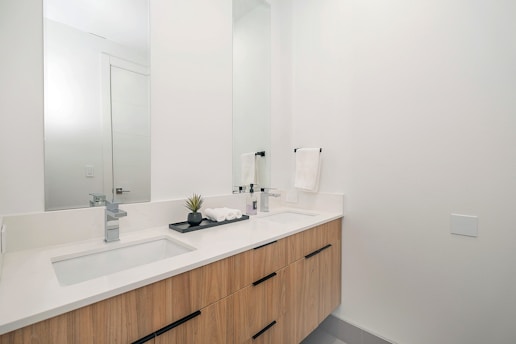 A modern bathroom with a sleek white countertop and light wood cabinetry. The countertop features a silver faucet, a decorative small plant, a tray with toiletries, and neatly folded towels. A large mirror extends above the sink area, reflecting the opposite wall. The walls are painted white, contributing to a clean and minimalist aesthetic.