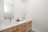 A modern bathroom with a sleek white countertop and light wood cabinetry. The countertop features a silver faucet, a decorative small plant, a tray with toiletries, and neatly folded towels. A large mirror extends above the sink area, reflecting the opposite wall. The walls are painted white, contributing to a clean and minimalist aesthetic.