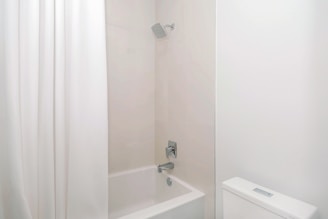 A minimalist bathroom setting featuring a white shower curtain, a bathtub, and a showerhead mounted on a tiled wall. The space is clean and brightly lit, with a focus on neutral tones.