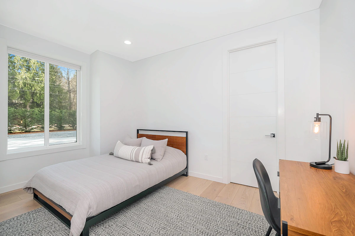 A bright student residence room with a single wood-framed bed, a wooden desk and a black accent chair beside a sunlit window looking out on trees