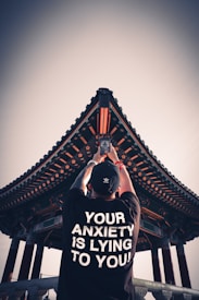 A person stands with their back to the camera, raising a smartphone, capturing a traditional architectural structure with intricate patterns and a triangular roof. The person is wearing a black shirt with the bold white text 'YOUR ANXIETY IS LYING TO YOU!' printed on the back.