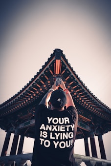 A person stands with their back to the camera, raising a smartphone, capturing a traditional architectural structure with intricate patterns and a triangular roof. The person is wearing a black shirt with the bold white text 'YOUR ANXIETY IS LYING TO YOU!' printed on the back.