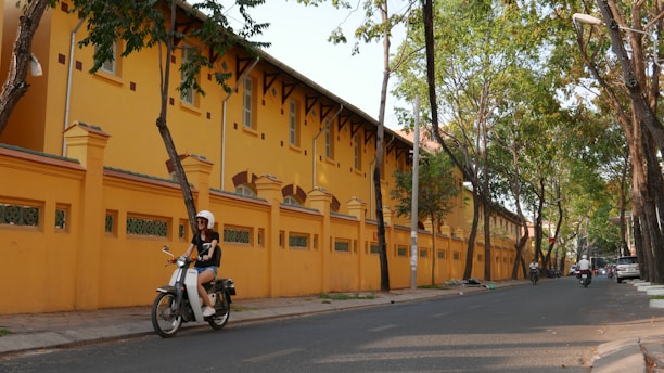 A person rides a scooter down a quiet street lined with tall trees and a long, two-story yellow building with architectural details. The road is mostly empty, and the atmosphere is calm and peaceful.