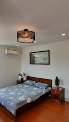A tidy bedroom featuring a double bed with blue bedding adorned with white clouds. The bed is flanked by two wooden nightstands, one with a vase of flowers and the other with a small potted plant. Above the bed, there is a framed painting on a white wall. The ceiling light has a black lace-like shade, and an air conditioning unit is mounted on the left wall.