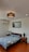 A tidy bedroom featuring a double bed with blue bedding adorned with white clouds. The bed is flanked by two wooden nightstands, one with a vase of flowers and the other with a small potted plant. Above the bed, there is a framed painting on a white wall. The ceiling light has a black lace-like shade, and an air conditioning unit is mounted on the left wall.
