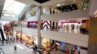 A multi-level shopping mall with various stores and people walking or shopping. The floors have a modern tiled pattern, and the upper levels have glass railings. Visible store signs include DAISO and other retail shops. There's also an escalator visible on the left side of the image.