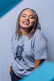 A person with a cheerful expression, wearing a light gray t-shirt with a black design and the words 'Royal Team.' The individual has brown braided hair and is standing against a plain backdrop.