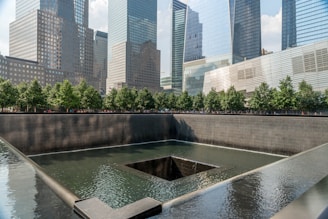 A reflective water feature with a dark square pool at its center is bordered by a low wall. In the background, there are tall modern skyscrapers with glass facades. Behind the pool, a line of green trees adds a touch of nature to the urban scene.