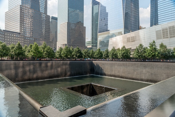 A reflective water feature with a dark square pool at its center is bordered by a low wall. In the background, there are tall modern skyscrapers with glass facades. Behind the pool, a line of green trees adds a touch of nature to the urban scene.