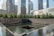 A reflective water feature with a dark square pool at its center is bordered by a low wall. In the background, there are tall modern skyscrapers with glass facades. Behind the pool, a line of green trees adds a touch of nature to the urban scene.