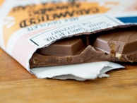 A partially unwrapped bar of chocolate lies on a wooden surface, revealing pieces of nuts embedded within the chocolate. The packaging is slightly torn and features text with a stylish design.