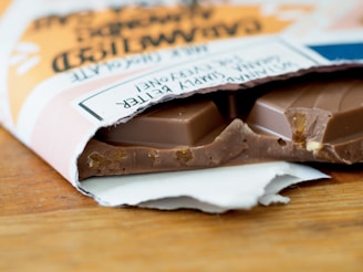 A partially unwrapped bar of chocolate lies on a wooden surface, revealing pieces of nuts embedded within the chocolate. The packaging is slightly torn and features text with a stylish design.