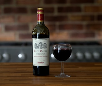 A bottle of Calvet Prestige Bordeaux wine and a filled wine glass are placed on a wooden table. The background features a blurred brick wall and part of a kitchen appliance.