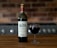 A bottle of Calvet Prestige Bordeaux wine and a filled wine glass are placed on a wooden table. The background features a blurred brick wall and part of a kitchen appliance.
