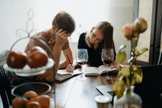 Two people are seated at a wooden table, engaged in writing or studying together with open notebooks and pens. There are two glasses of red wine in front of them. A laptop and a metal fruit basket with apples are also on the table. A vase with a rose is in the foreground, adding a touch of warmth to the setting.