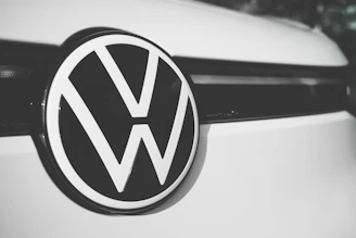 A close-up of a Volkswagen logo centered on the front of a vehicle, displaying the iconic VW letters in a bold, black and white design against a smooth, clean surface.