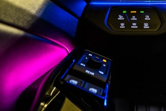 A car's interior control panel is illuminated with vivid colors, featuring buttons and switches for various functionalities like front and rear control, temperature settings, and automatic mode. The lighting creates a striking contrast against the dark dashboard.