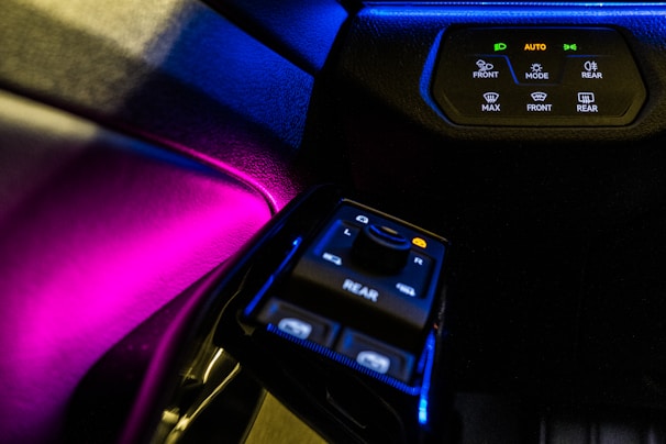 A car's interior control panel is illuminated with vivid colors, featuring buttons and switches for various functionalities like front and rear control, temperature settings, and automatic mode. The lighting creates a striking contrast against the dark dashboard.