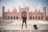 A man is standing confidently in front of an impressive red sandstone mosque with large domes and minarets. The architecture is intricate and grand, showcasing detailed carvings and symmetrical design. A backpack is placed on the ground next to the man.