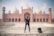 A man is standing confidently in front of an impressive red sandstone mosque with large domes and minarets. The architecture is intricate and grand, showcasing detailed carvings and symmetrical design. A backpack is placed on the ground next to the man.