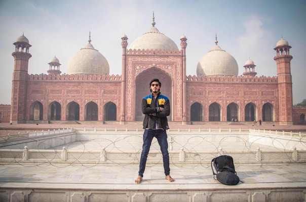 A man is standing confidently in front of an impressive red sandstone mosque with large domes and minarets. The architecture is intricate and grand, showcasing detailed carvings and symmetrical design. A backpack is placed on the ground next to the man.