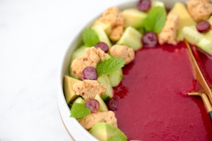 A bowl filled with a thick red smoothie-like substance, garnished with green apple chunks, granola, grape slices, and fresh mint leaves. A wooden spoon is placed on the side.