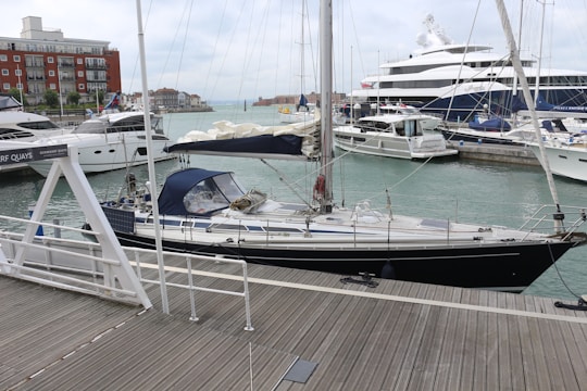 A marina populated with several boats and yachts. A sailboat with dark hull and cream-colored deck is docked close to a wooden platform. Behind it, larger luxury yachts are anchored. In the background, a row of buildings lines the waterfront under a cloudy sky.