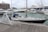 A marina populated with several boats and yachts. A sailboat with dark hull and cream-colored deck is docked close to a wooden platform. Behind it, larger luxury yachts are anchored. In the background, a row of buildings lines the waterfront under a cloudy sky.