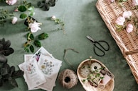 A collection of gardening items and flowers laid out on a green surface. There are various plants and roses scattered around, some in a wicker basket. Seed packets depicting different kinds of flowers, a ball of twine, and gardening shears are also visible. The scene is organized yet has a sense of natural flow.