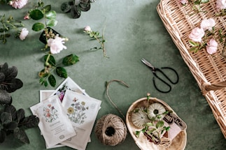 A collection of gardening items and flowers laid out on a green surface. There are various plants and roses scattered around, some in a wicker basket. Seed packets depicting different kinds of flowers, a ball of twine, and gardening shears are also visible. The scene is organized yet has a sense of natural flow.