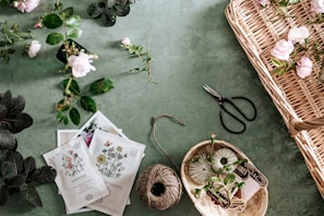 A collection of gardening items and flowers laid out on a green surface. There are various plants and roses scattered around, some in a wicker basket. Seed packets depicting different kinds of flowers, a ball of twine, and gardening shears are also visible. The scene is organized yet has a sense of natural flow.