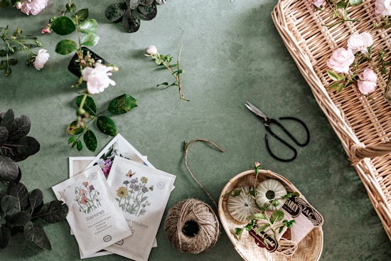 A collection of gardening items and flowers laid out on a green surface. There are various plants and roses scattered around, some in a wicker basket. Seed packets depicting different kinds of flowers, a ball of twine, and gardening shears are also visible. The scene is organized yet has a sense of natural flow.