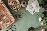 A green surface is scattered with gardening supplies, including a wicker basket with pink flowers, twine, pruning shears, seed packets, and a small basket containing various threads and yarns. Several leafy branches and roses are also present.