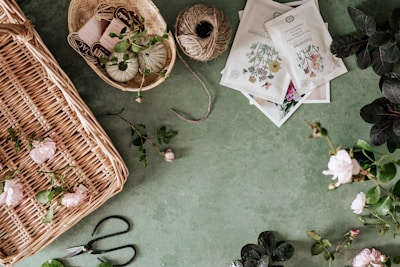 A green surface is scattered with gardening supplies, including a wicker basket with pink flowers, twine, pruning shears, seed packets, and a small basket containing various threads and yarns. Several leafy branches and roses are also present.