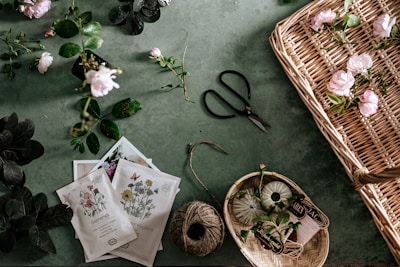 A tabletop is arranged with various gardening and floral items. Scattered pink roses and green leaves contrast against a muted green background. Seed packets are displayed alongside gardening scissors, a ball of twine, and a wicker basket partially filled with flowers. Smaller baskets contain more gardening tools and supplies.