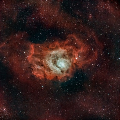 A vast cosmic scene featuring a bright nebula with a glowing core surrounded by swirling red and orange clouds. Countless stars are scattered throughout the dark expanse, adding to the depth and complexity of the scene.