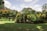 A lush garden featuring a row of vibrant green and orange-leaved bushes under a partly cloudy sky. The well-maintained grass lawn in the foreground is bordered by neatly shaped plants, indicating careful landscaping. The bright sunlight casts shadows of leaves and trees on the ground.