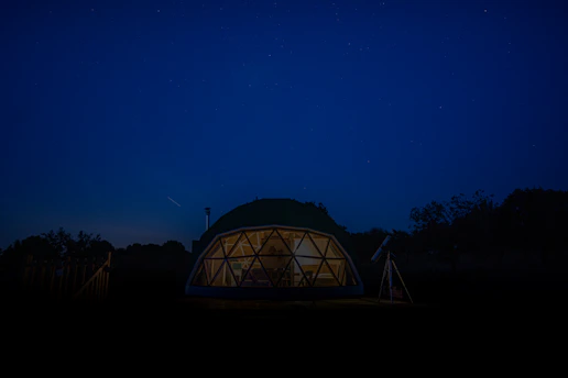 A geodesic dome structure is illuminated from within against a night sky, surrounded by dark silhouettes of trees. A telescope stands on the right side, hinting at stargazing activities.