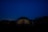 A geodesic dome structure is illuminated from within against a night sky, surrounded by dark silhouettes of trees. A telescope stands on the right side, hinting at stargazing activities.