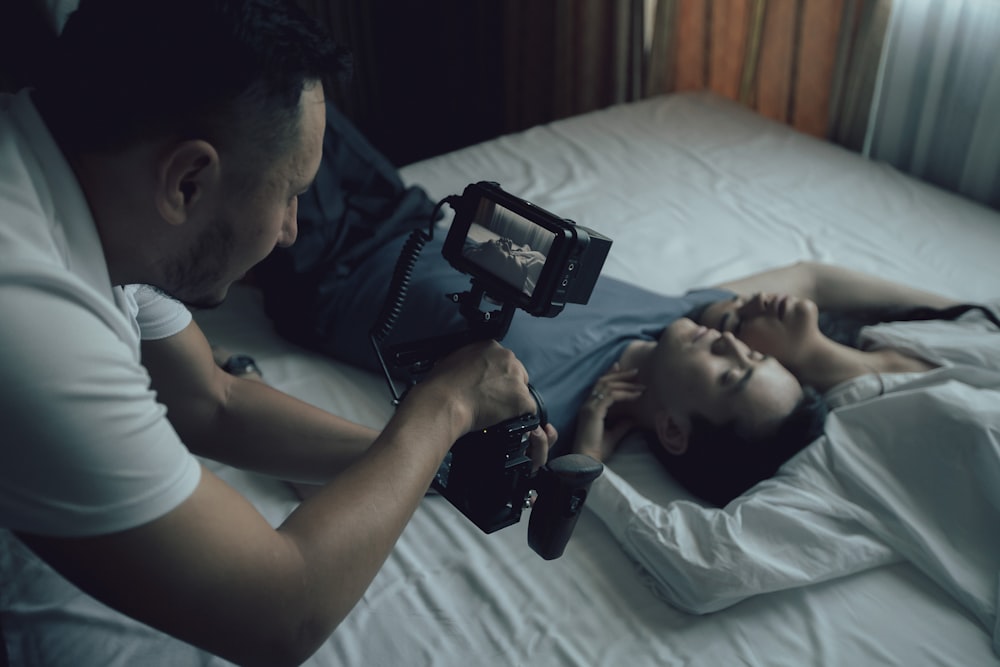 man in white tank top lying on bed holding black smartphone