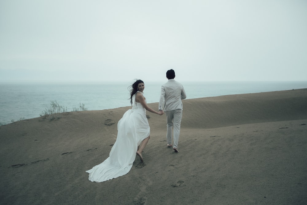 man and woman in white wedding dress walking on brown sand near body of water during
