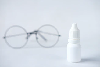 A small white plastic bottle with a dropper cap is placed in the foreground, while a pair of round eyeglasses is blurred in the background. The scene appears minimalist with a clean white background.