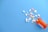 A variety of colored pills and tablets are scattered on a light blue surface, originating from an open orange prescription bottle lying on its side. The pills include different shapes and colors such as white, yellow, pink, and green.