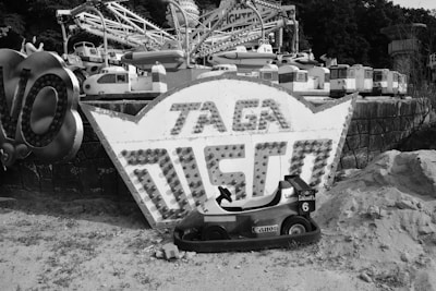 A vintage amusement park with a collection of small arcade ride cars placed on a dusty ground. A large retro sign with bulb lights displays the words 'Taga Disco'. The background shows an old, possibly abandoned, merry-go-round with several small car and train models.