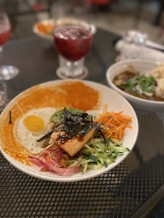 A colorful dish featuring a perfectly cooked salmon fillet atop a bed of shredded vegetables including carrots, cucumbers, and pickled onions, with a sunny-side-up egg nestled on one side. The dish is garnished with seaweed strips and sesame seeds, surrounded by a vibrant sauce. In the background, a glass of iced berry drink and another bowl with what appears to be a hearty soup with herbs are visible.